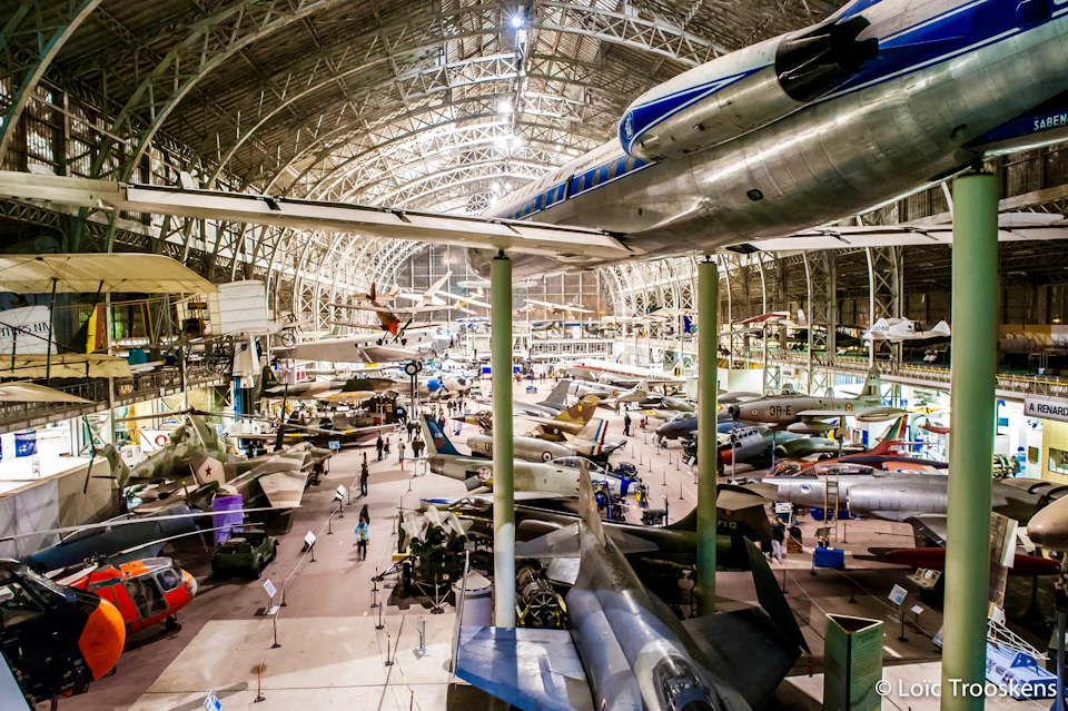 A large indoor aviation museum with various aircraft displayed on the floor and suspended from the ceiling, people walking around.