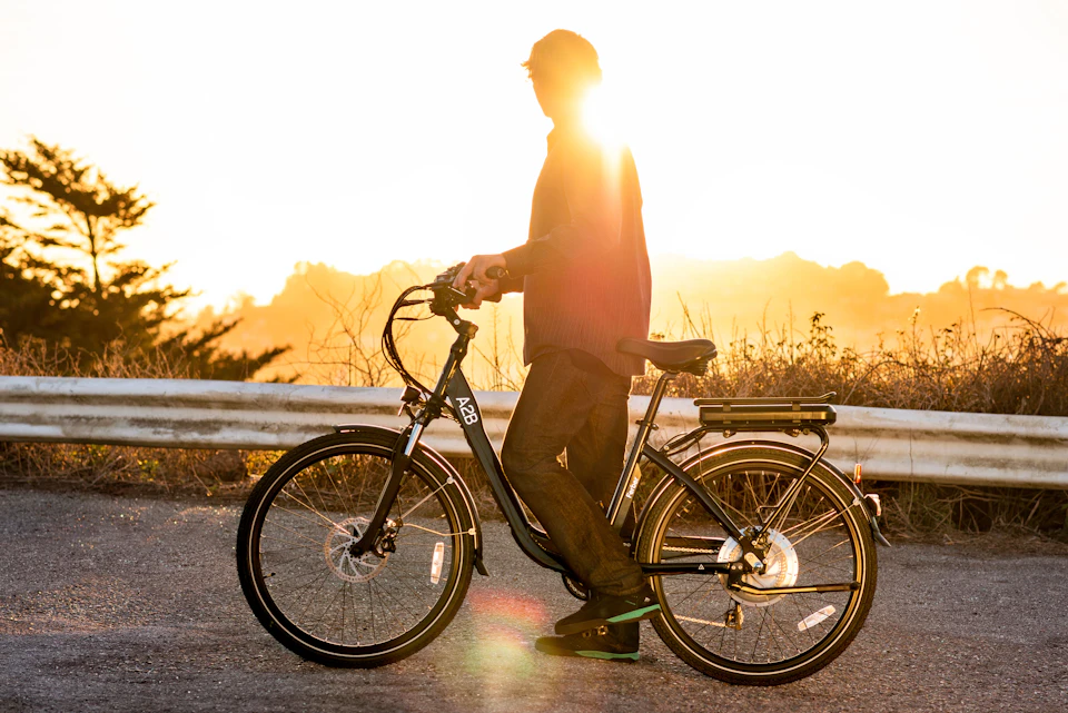 A person stands beside a black electric bicycle on a road, with a bright sunrise in the background and foliage along the guardrail.
