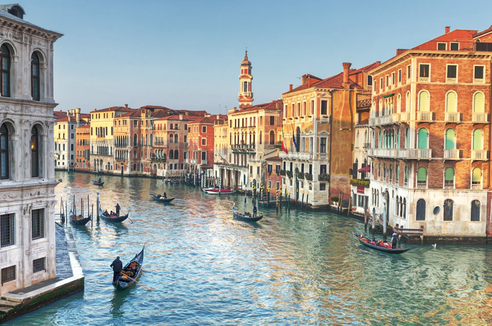 Gondolas on a Venetian canal with historic buildings and a tall bell tower lining the water under a clear sky.