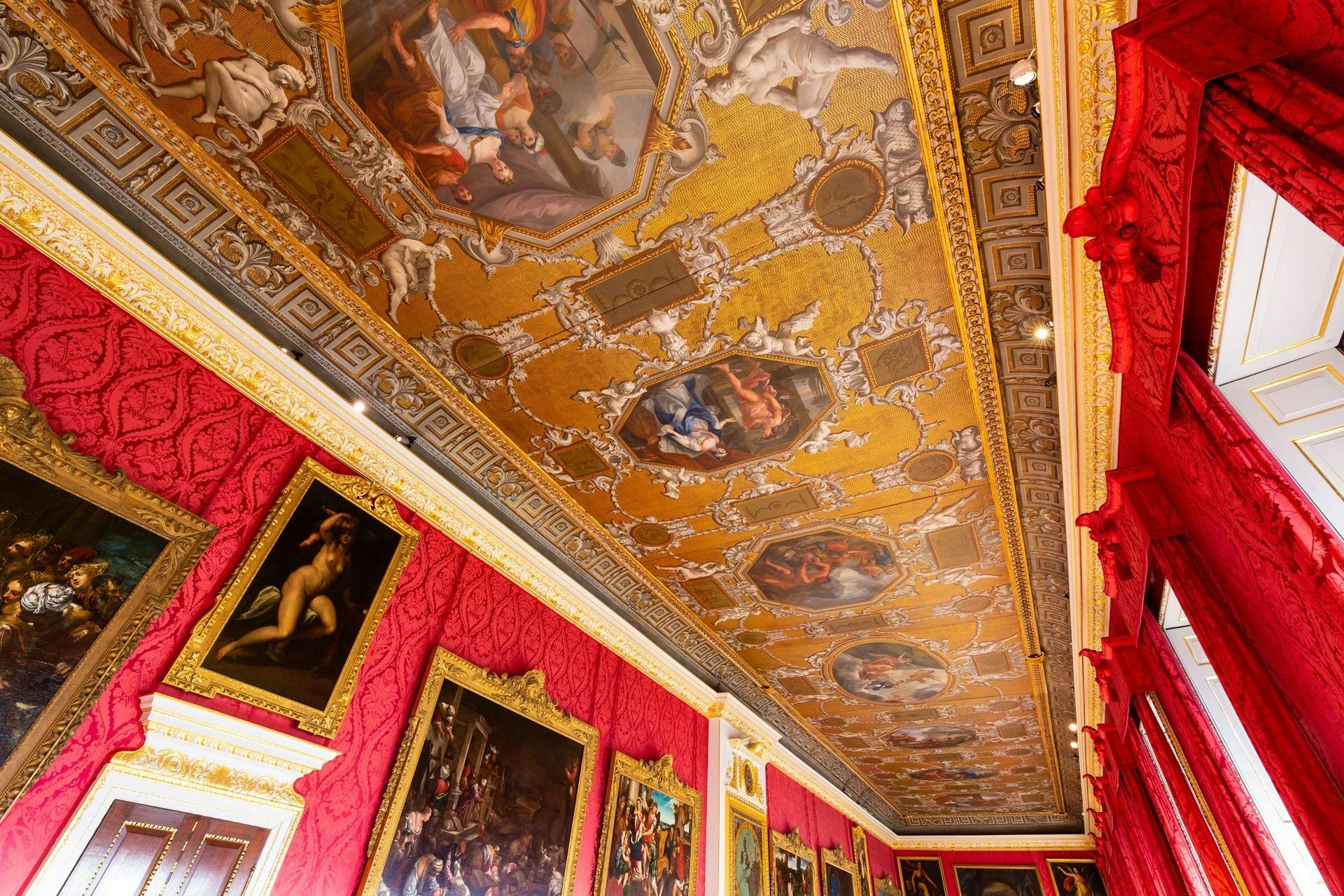 Ornate ceiling with intricate frescoes and gold detailing, surrounded by red walls adorned with framed paintings in a grand room.