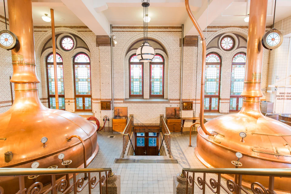 A brewery interior featuring copper brewing tanks and stained glass windows in a well-lit room with tiled flooring.