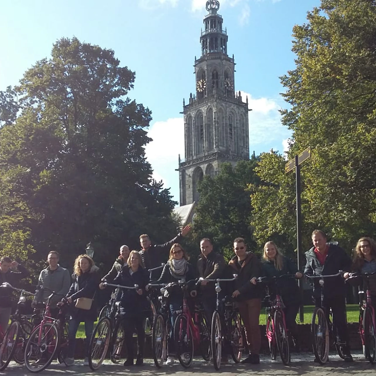 A group of people standing with bicycles in front of a tall, historic church tower surrounded by trees under a clear sky.