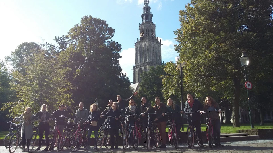 A group of people standing with bicycles in front of a tall, historic church tower surrounded by trees under a clear sky.