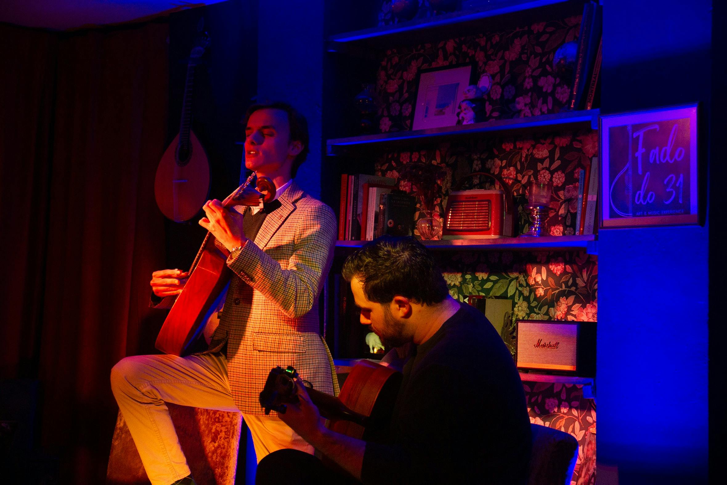 Δύο μουσικοί, ο ένας όρθιος με βιολί και ο άλλος καθιστός με κιθάρα, δίνουν παράσταση σε μια ζεστά φωτισμένη αίθουσα με μουσικά όργανα και βιβλία.