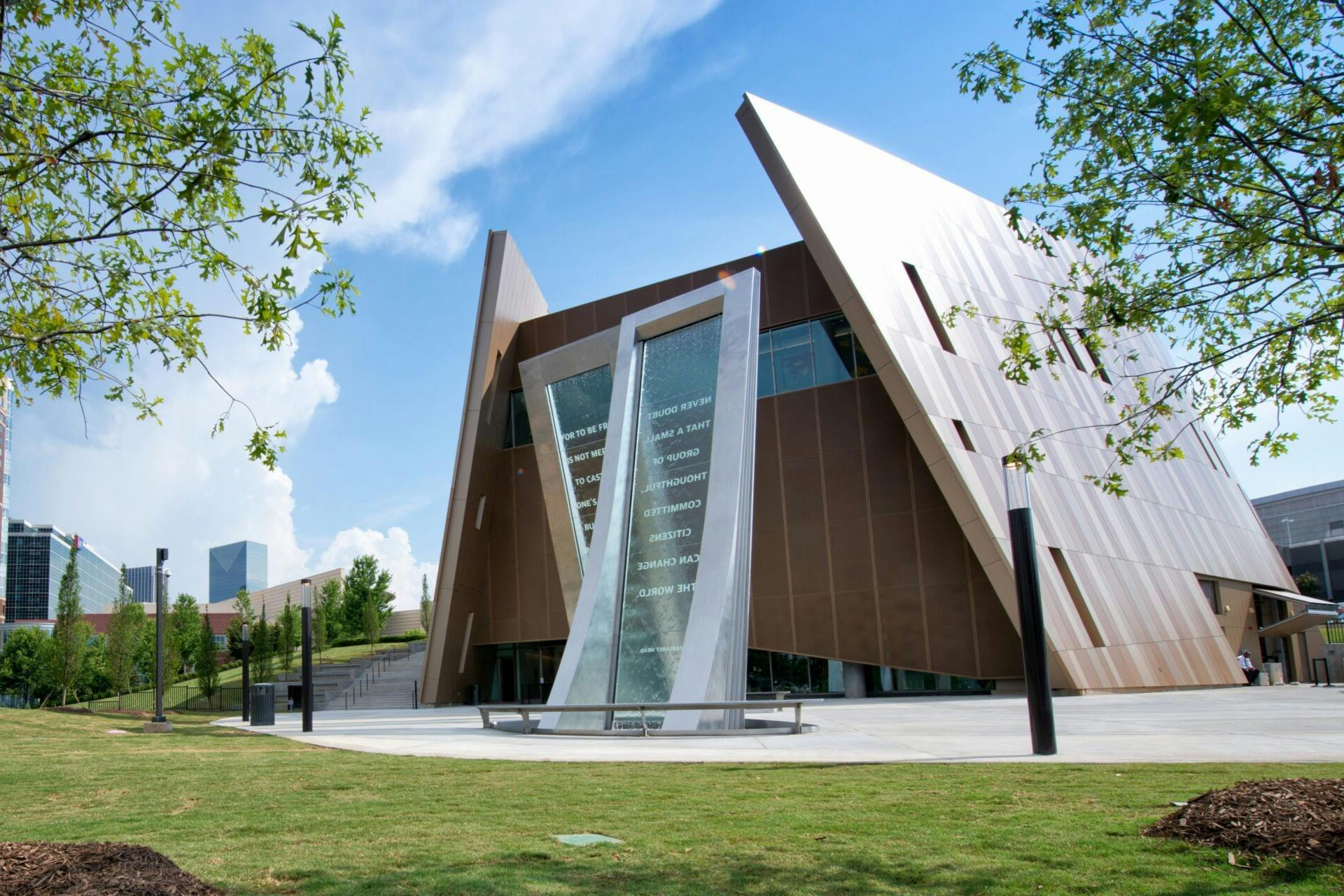 Um edifício moderno com um design angular e um monumento com uma inscrição em vidro em primeiro plano, rodeado de relva e árvores.