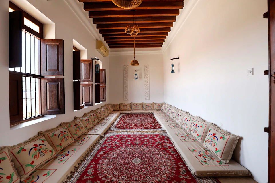 A long room with wooden ceiling beams, embroidered floor seating, red patterned rugs, and wooden shutters on the windows.
