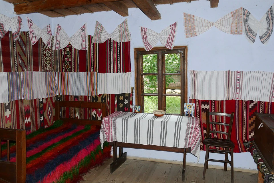 A rustic room with colorful woven textiles on the walls and furniture, a wooden table with a striped cloth, and a window showing greenery outside.