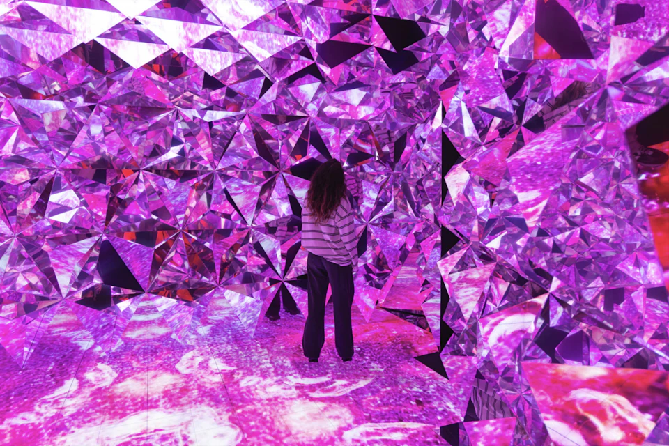 A person stands in a room filled with reflective, faceted surfaces illuminated by pink and purple lights.