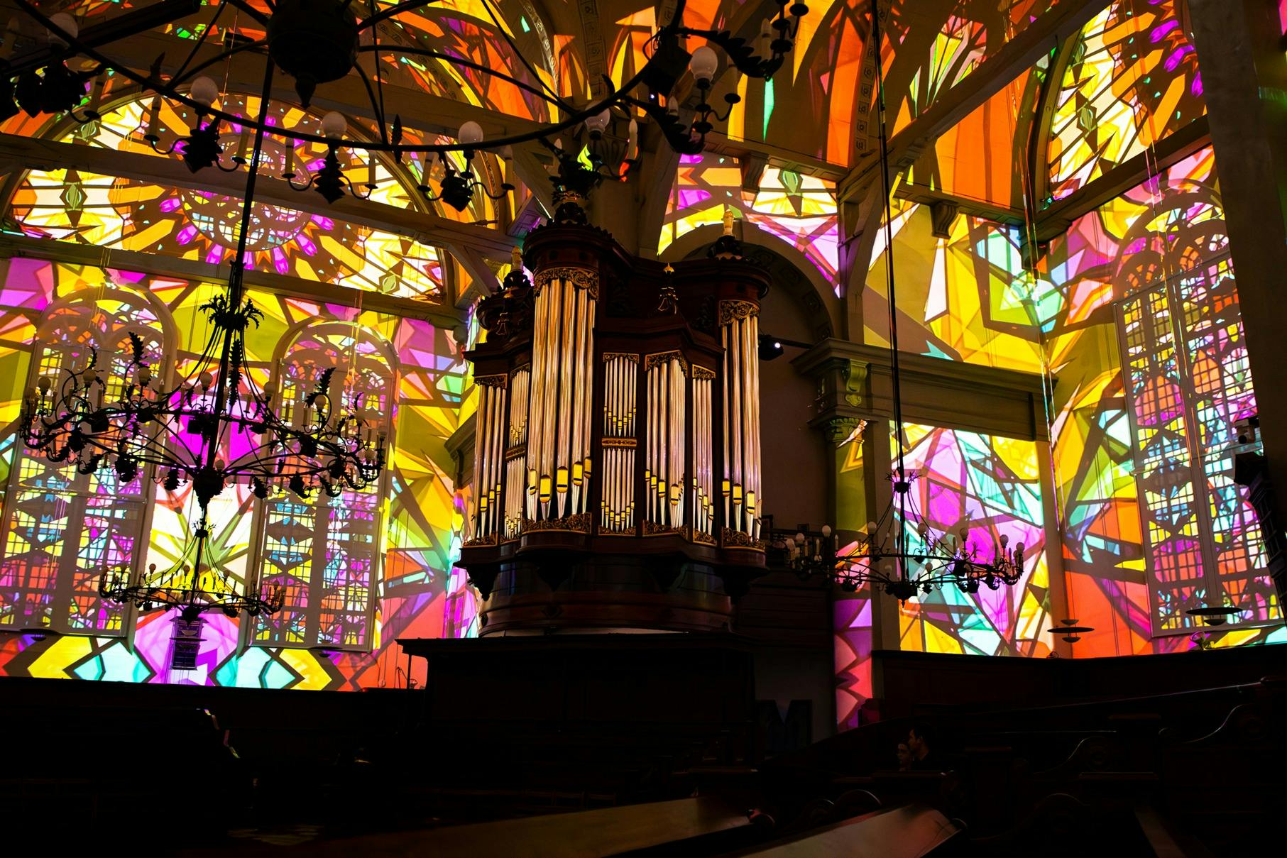 教会内にある大きなパイプオルガンで、ステンドグラスの窓が照らされており、壁や天井にはカラフルな光のパターンが描かれています。