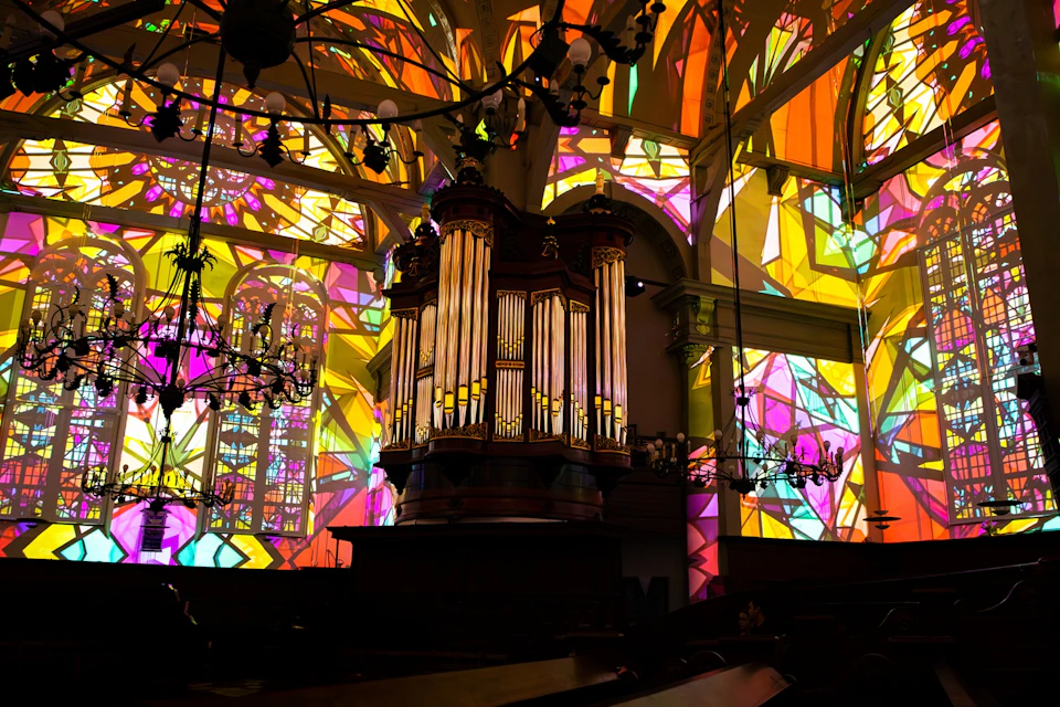 A large pipe organ inside a church with illuminated stained glass windows casting colourful light patterns on the walls and ceiling.