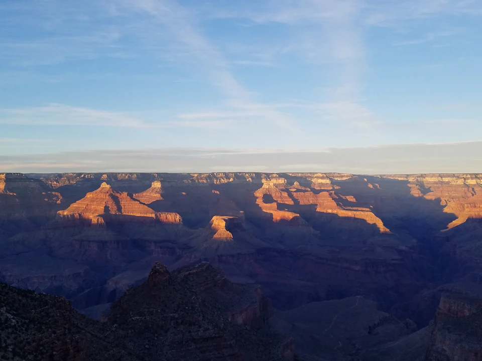 The image shows a vast landscape of the Grand Canyon at sunset with shadows highlighting the rugged terrain under a clear, blue sky.