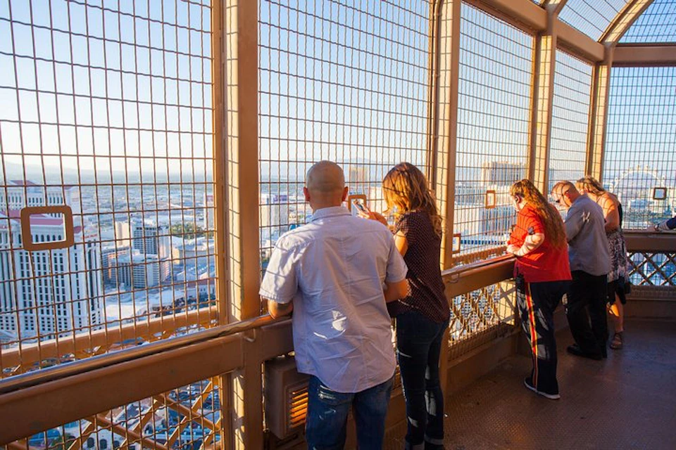 People standing on an observation deck behind a protective wire fence, overlooking a cityscape at sunset.