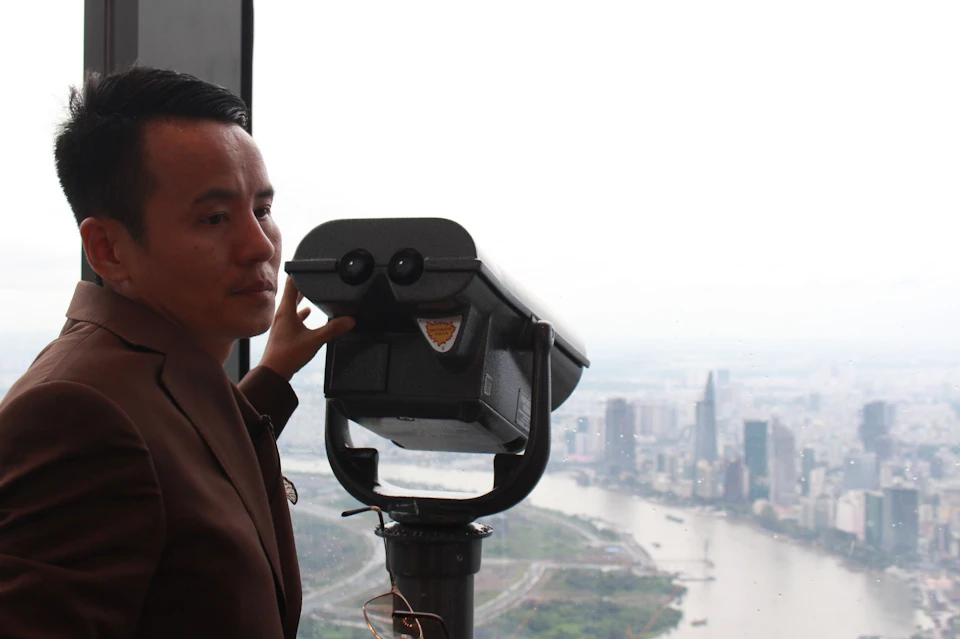 A person in a brown jacket looks through a binocular viewer in a high-rise building with a cityscape view in the background.