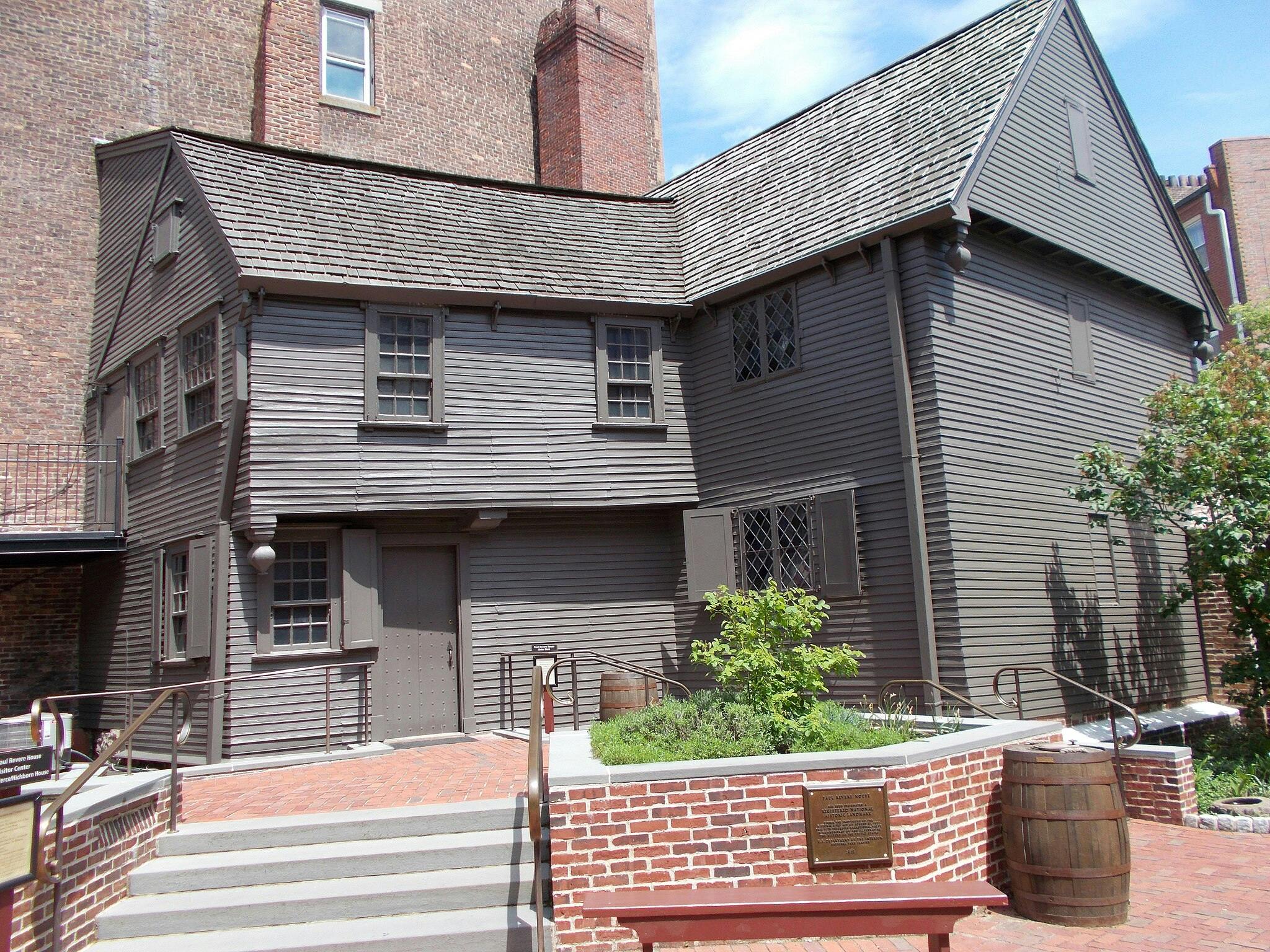 A two-story, wooden colonial house with a brick chimney, surrounded by a brick walkway, stairs, and a garden with a bench.