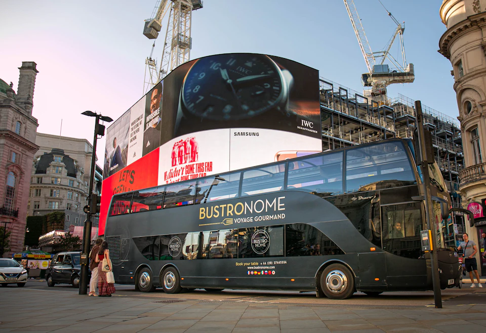 A black double-decker bus labeled "Bustronome" is parked near large digital billboards in an urban setting with cranes in the background.
