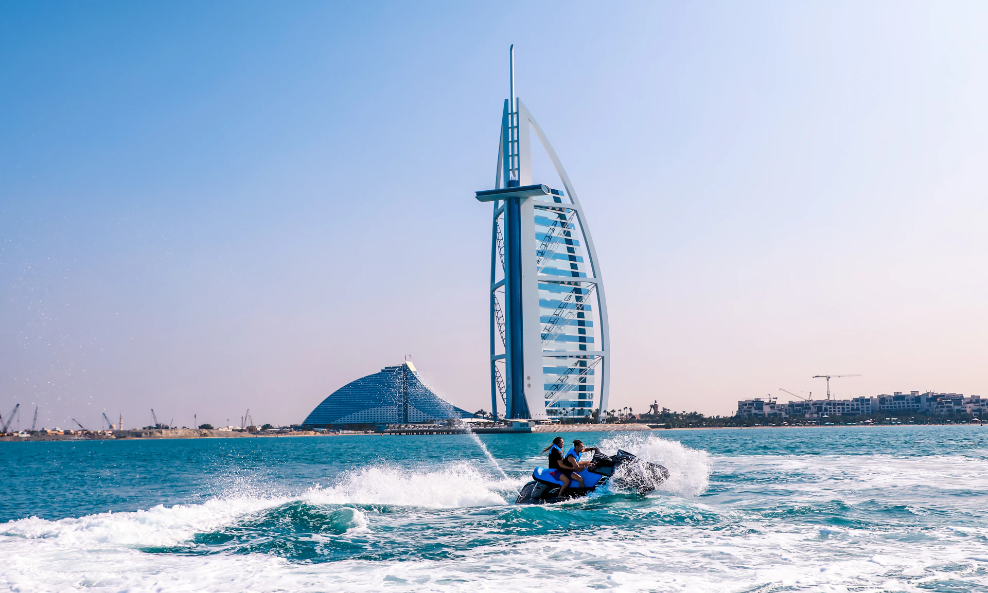 Two people ride a jet ski on blue waters with the Burj Al Arab hotel and another curved building in the background on a clear day.