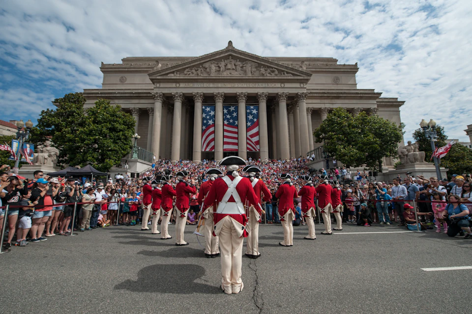 A group of performers in red uniforms stand before a crowd in front of a neoclassical building draped with a large U.S. flag.