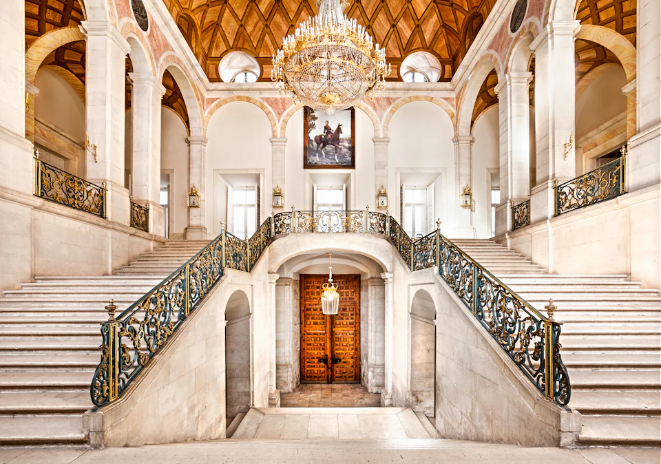 Grand marble staircase with ornate railings, a large chandelier, arched windows, and a wooden door beneath a painting.