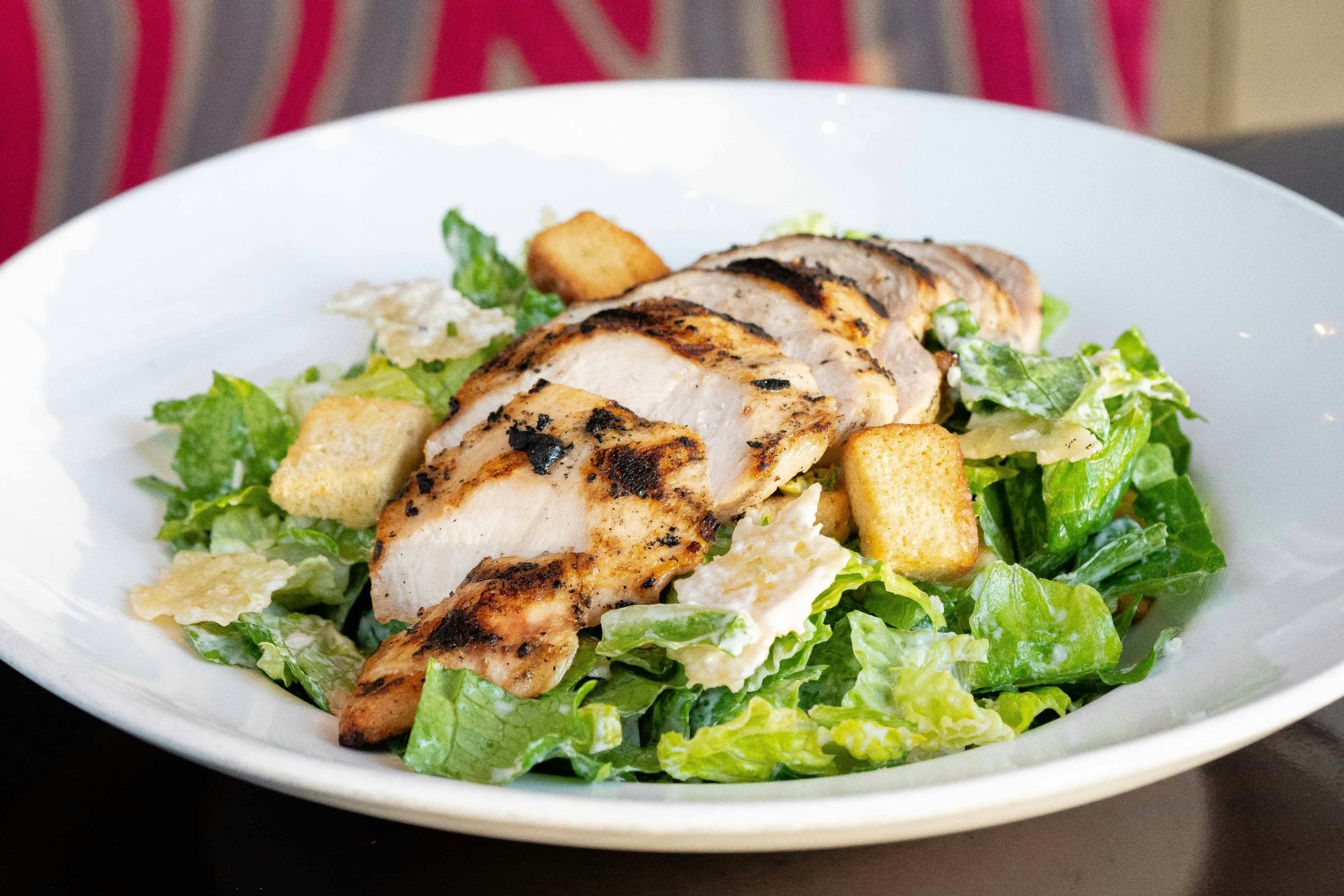 Grilled chicken slices on a Caesar salad with croutons and romaine lettuce in a white bowl.