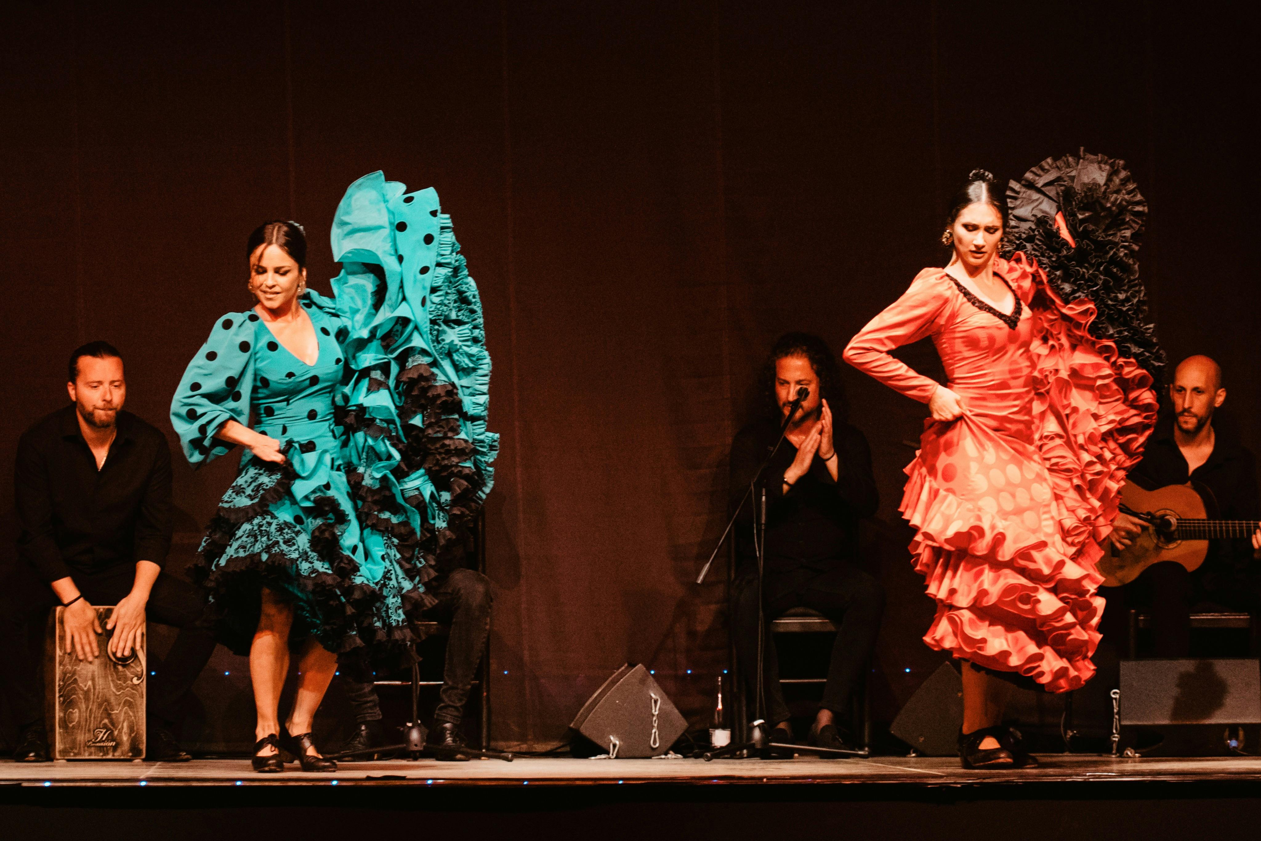 Deux danseuses de flamenco vêtues de robes vibrantes à volants se produisent sur scène avec trois musiciens jouant de la guitare, de la boîte à rythmes et tapant des mains.