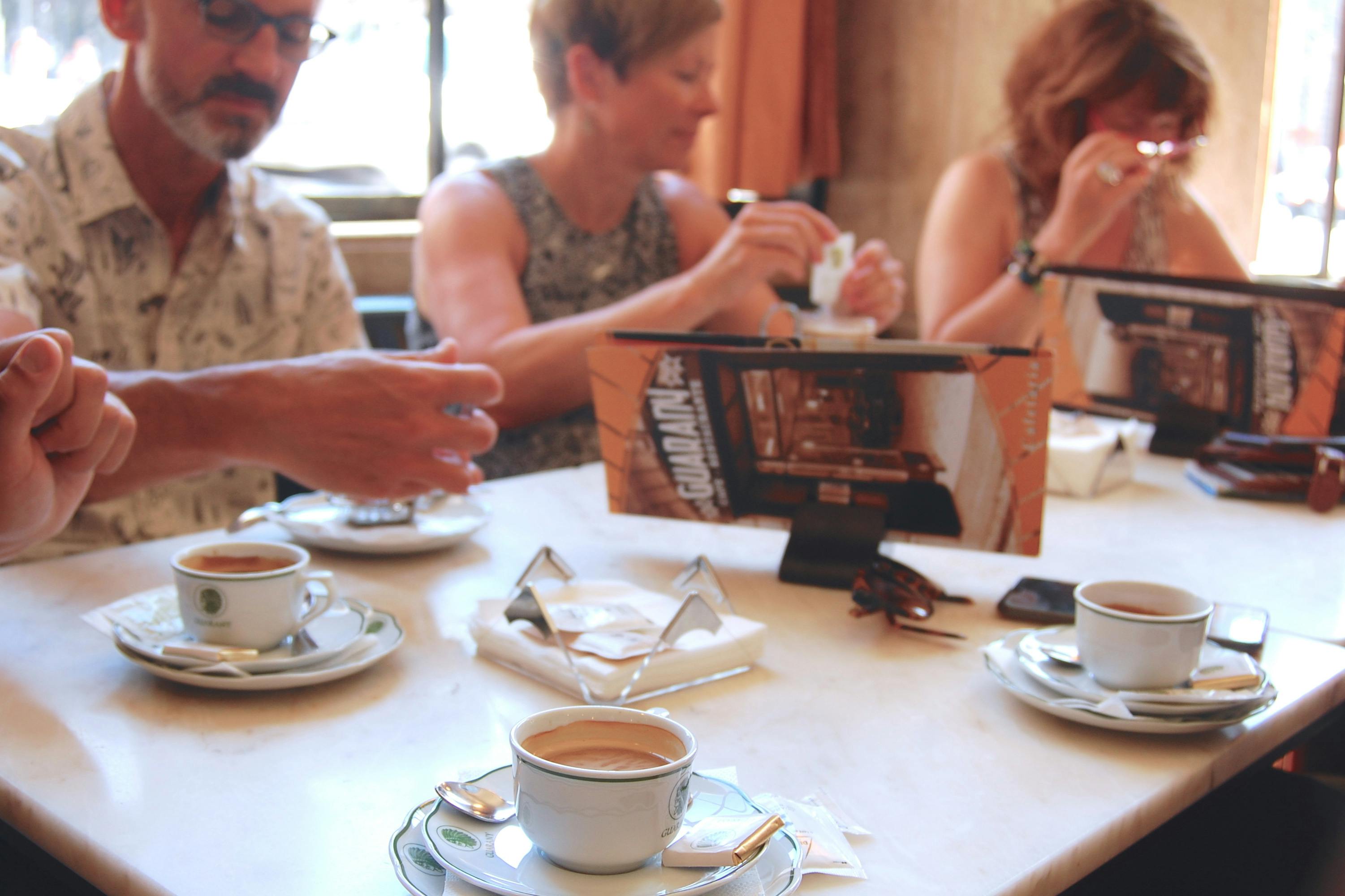 Τρία άτομα κάθονται σε ένα τραπέζι και απολαμβάνουν καφέ. Ένα μικρό μενού τοποθετείται όρθιο στο κέντρο του τραπεζιού.