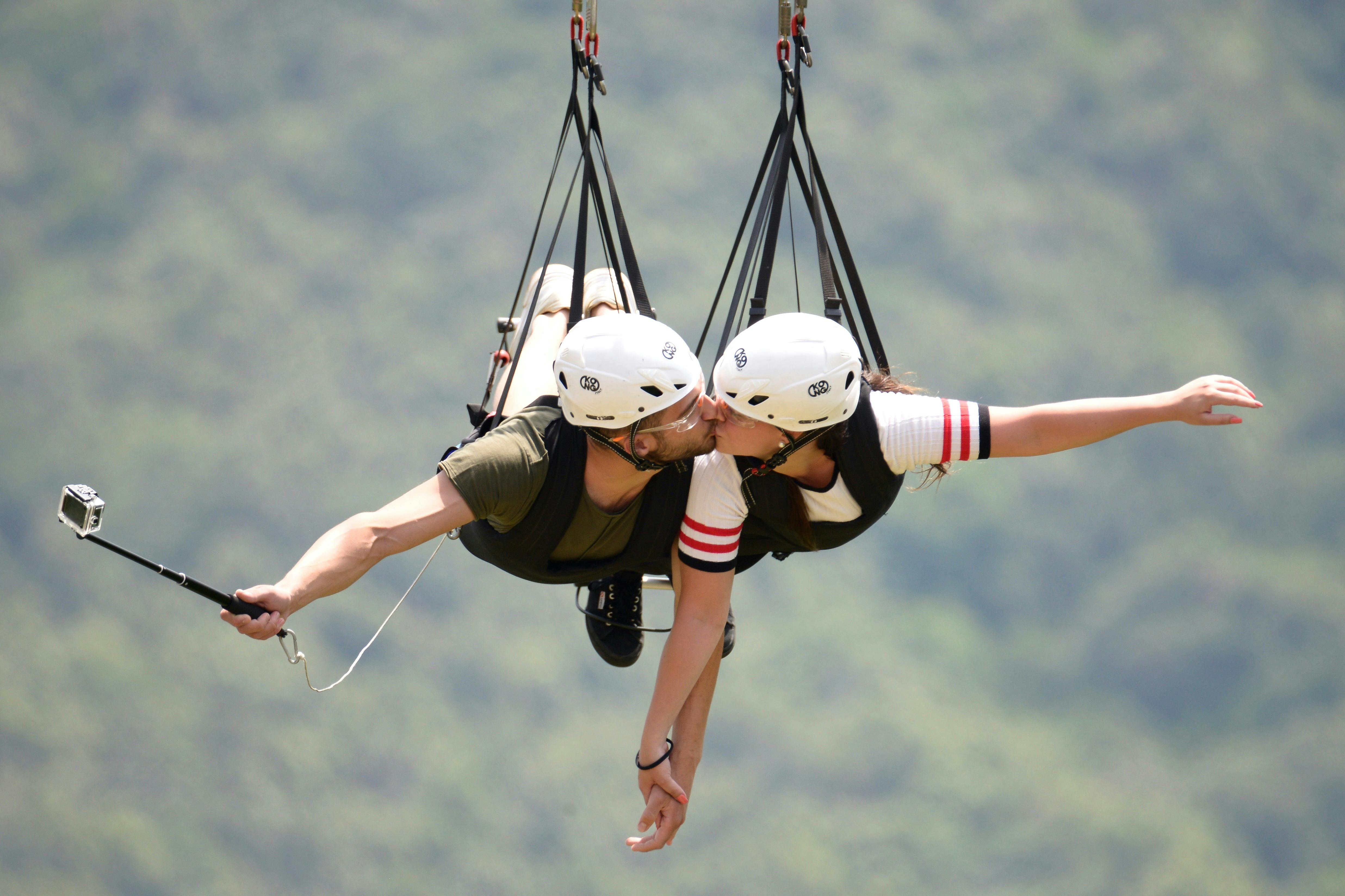 一对戴着头盔的夫妇吊在高空滑索上亲吻，背景是模糊的绿色景观。