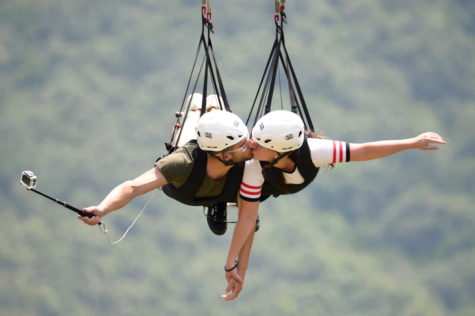 A couple wearing helmets kiss while suspended in a zipline harness with a blurred green landscape in the background.