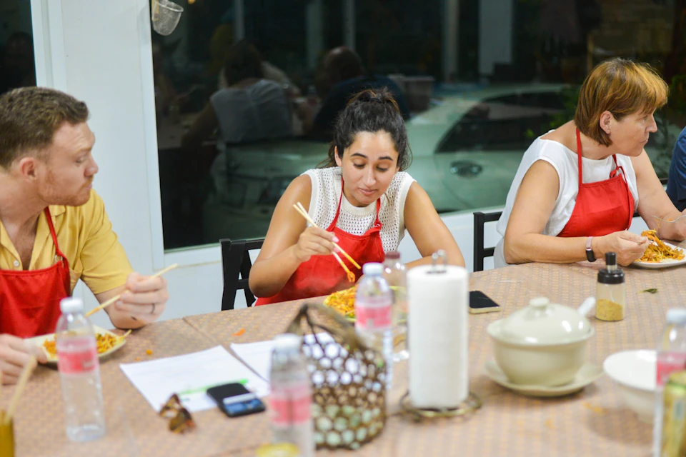 A woman in a red apron eats with chopsticks at a table set with dishes, a paper towel roll, and bottles.
