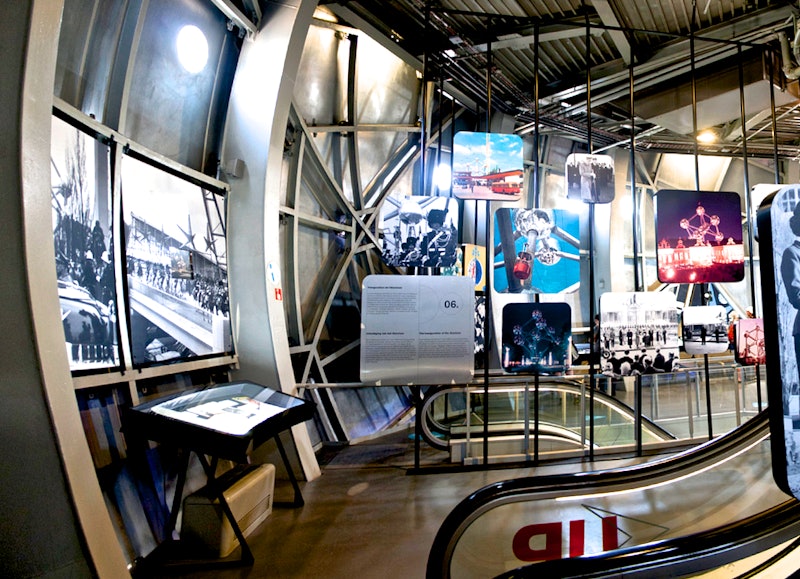 Eksponat wewnętrzny z różnymi historycznymi fotografiami i panelami wyświetlanymi na metalowych ramach, w pobliżu schodów ruchomych.