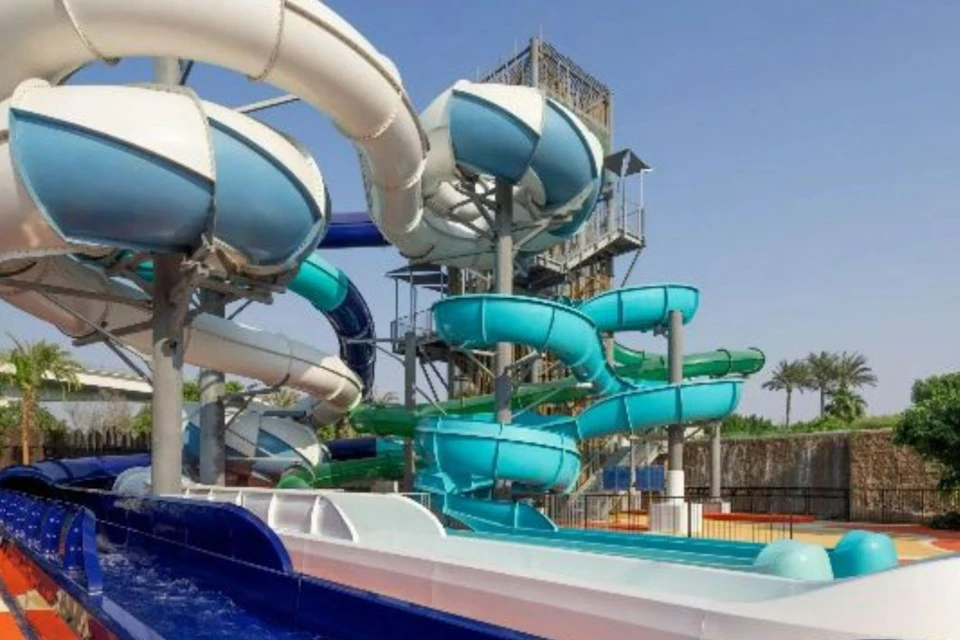 A water slide park featuring multiple twisting slides in blue and green, with a tall platform against a clear blue sky.