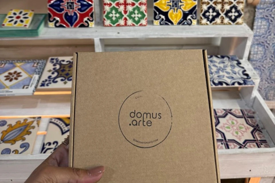A hand holding a cardboard box labeled "domus arte" in front of a display of colorful decorative tiles on shelves.