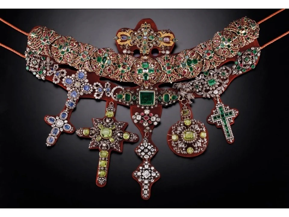 Intricately designed jewelry with green, blue, and red gemstones featuring crosses and a central green stone on a black background.