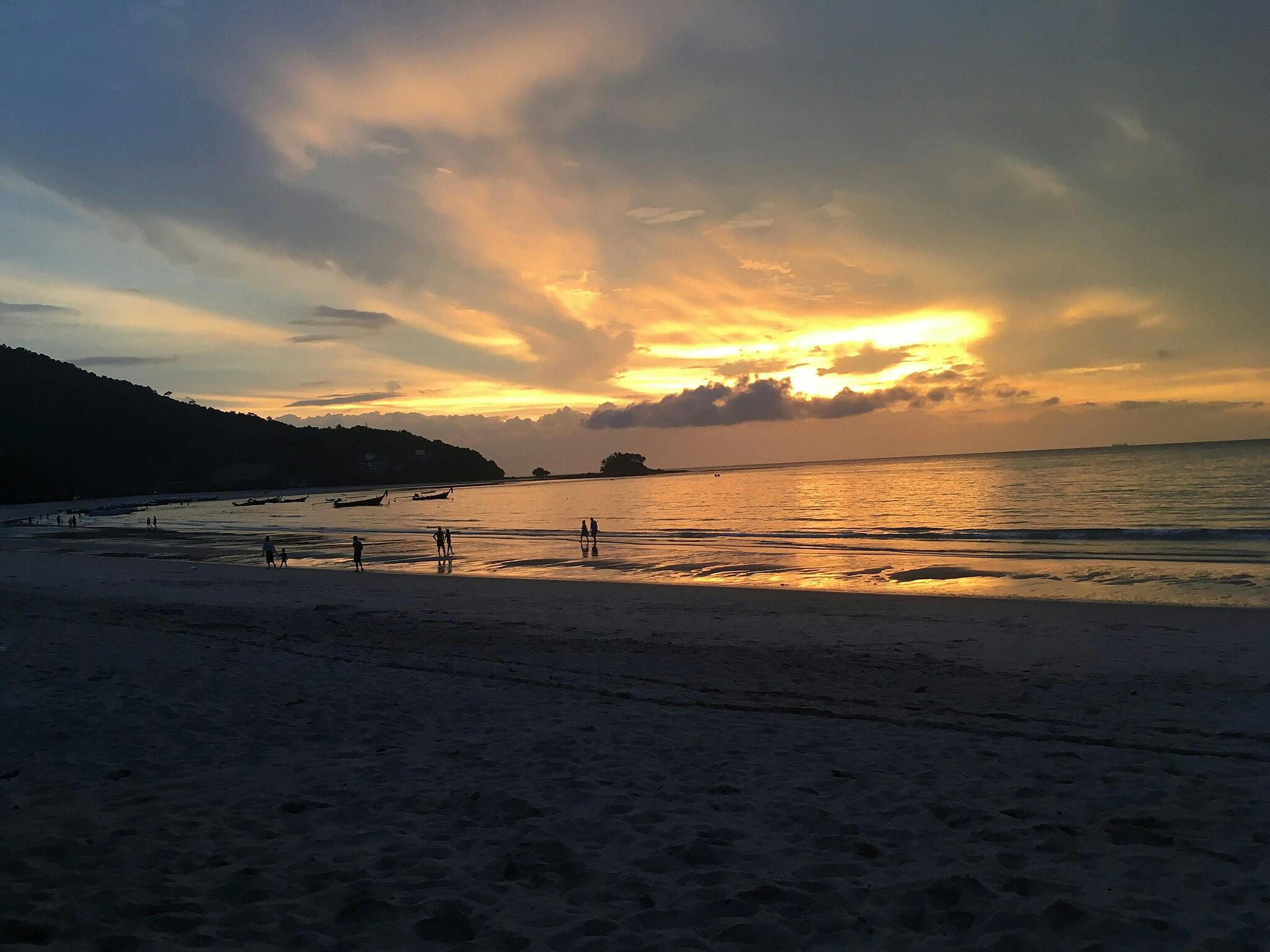 People walking along a beach at sunset with small boats on the water and silhouetted hills in the background.