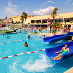 Fullsatt utomhusvattenpark med människor som simmar, leker och använder rutschkanor och fontäner under en solig himmel.