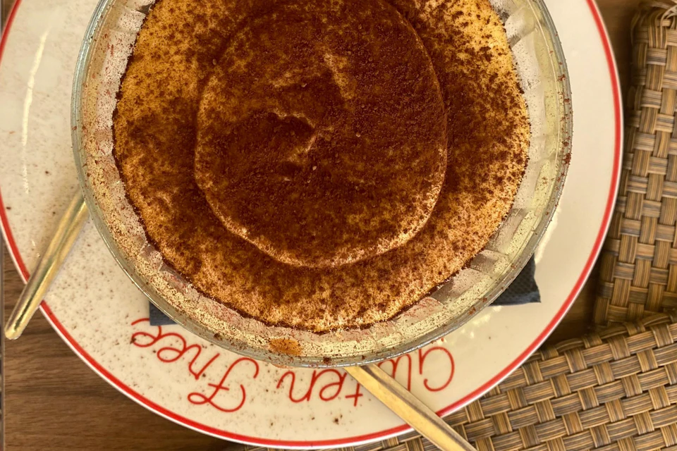 Tiramisu in a glass dish on a white plate with red text, placed on a woven table mat with a spoon on the side.