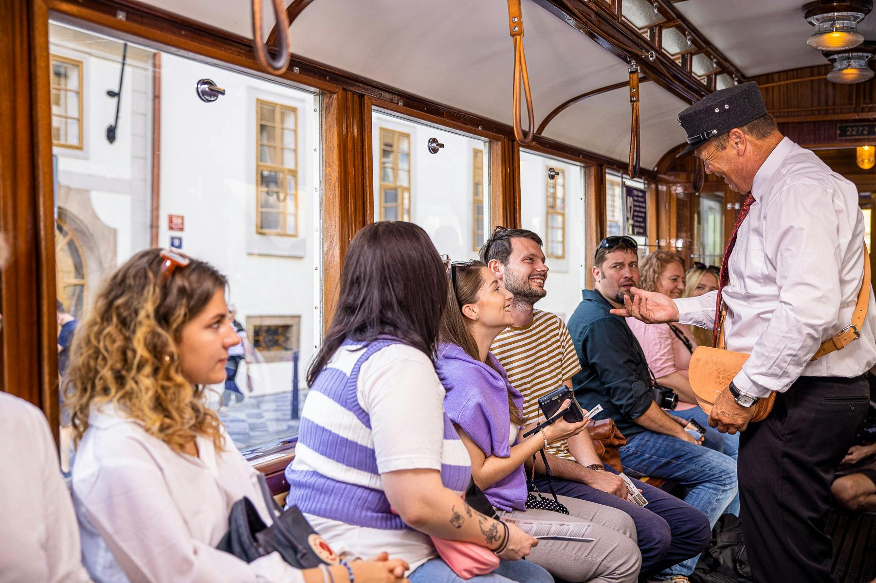 Μια ομάδα ανθρώπων κάθισε μέσα σε ένα τραμ, κοιτάζοντας μεγάλα παράθυρα σε έναν δρόμο με κτίρια. Ένα άτομο κρατά μια κάμερα.