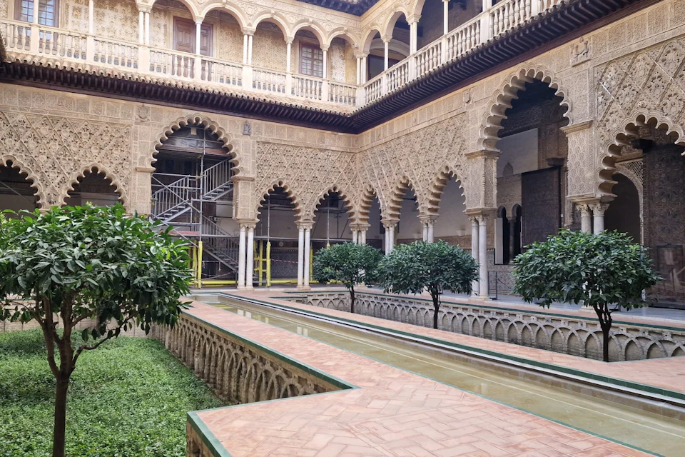 Courtyard with intricate arches, a central water feature, surrounding greenery, and visible scaffolding in one corner.