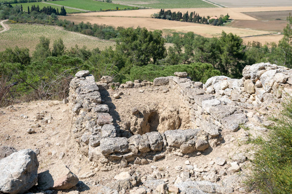 Ruins of a small stone structure with a hole in the center, surrounded by a rural landscape with fields and trees.