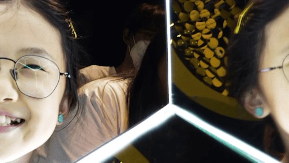 A smiling girl with glasses is reflected multiple times in a hexagonal mirror pattern, creating a kaleidoscopic effect.