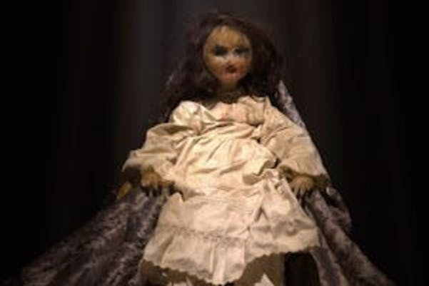 En docka med långt mörkt hår iklädd en vit klänning, sittande på en tygbakgrund under dramatisk belysning.