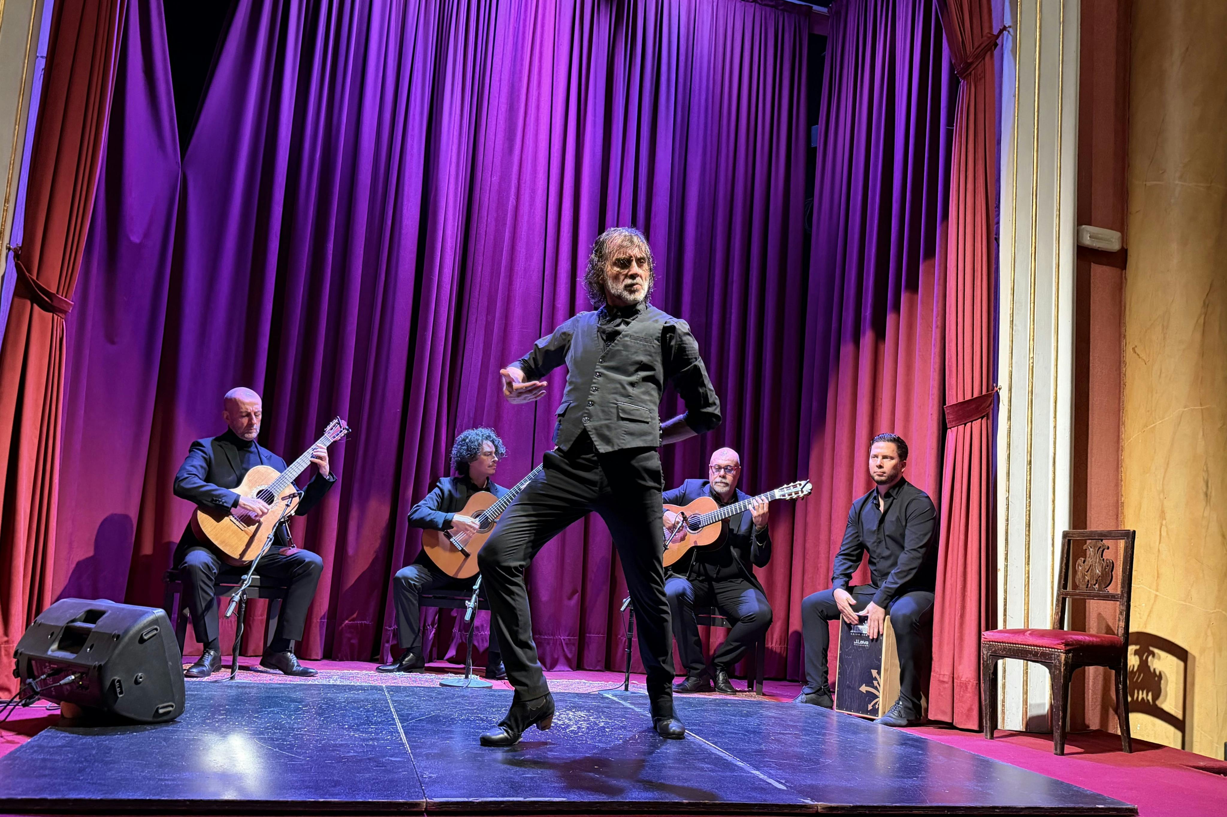 Un danseur de flamenco se produit sur scène avec quatre musiciens assis derrière lui, jouant de la guitare et du cajón. L'arrière-plan est constitué de rideaux rouges.