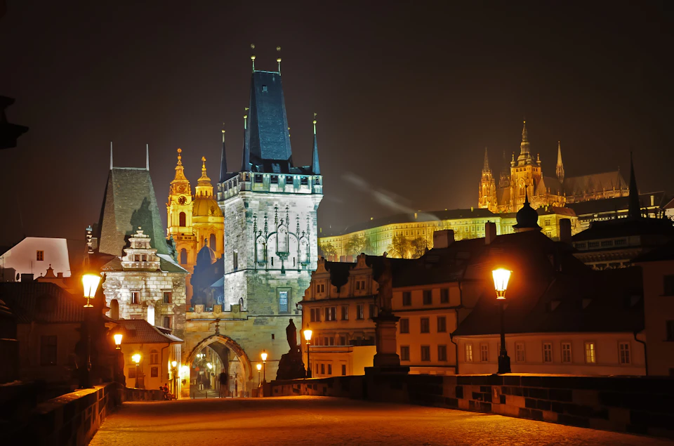 Historic cityscape at night featuring a lit bridge, a stone gate tower, and illuminated buildings in the background.