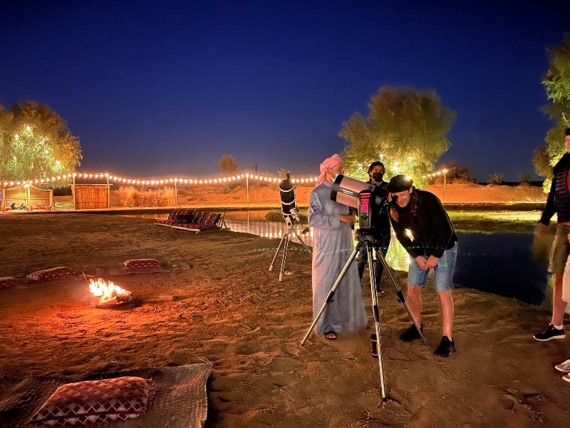 夜、小さなキャンプファイヤーの近くにある望遠鏡を見ている2人の人物は、ひもの光と木に囲まれています。