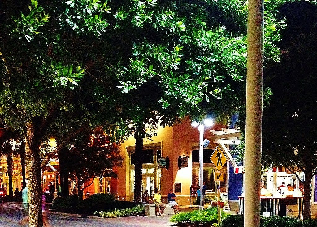 Scène de rue nocturne avec des arbres feuillus, des devantures de magasins éclairées par des lumières. Les gens s'assoient et se promènent, et un panneau de signalisation de passage pour piétons est visible.