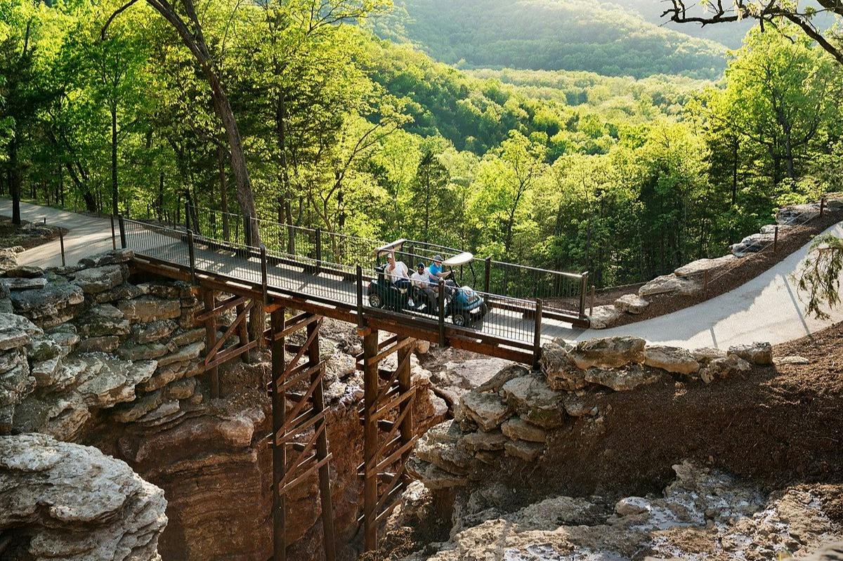 A small group of people riding a golf cart on a bridge over a rocky ravine, surrounded by a forested landscape.