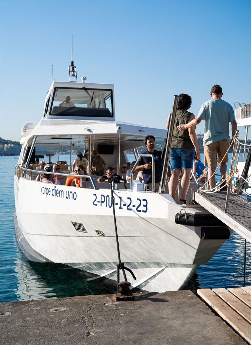 Μια ομάδα ανθρώπων επιβιβάζεται σε μια λευκή βάρκα με το όνομα «Carpe Diem Uno» μια ηλιόλουστη μέρα, αγκυροβολημένη σε μια αποβάθρα.