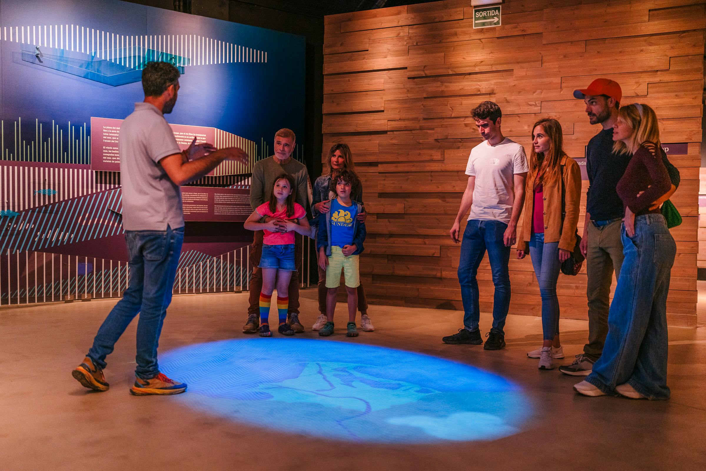 Μια ομάδα ανθρώπων στέκεται σε μια αίθουσα εκθεμάτων με μπλε και ξύλινους τοίχους και μια προβολή στο πάτωμα.