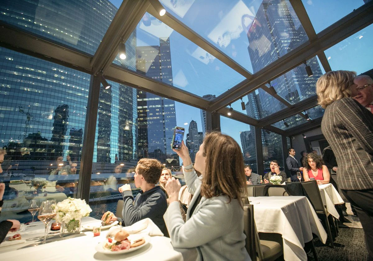 People dining in a glass-enclosed restaurant with a city skyline view at dusk; one person is taking a photo with a smartphone.
