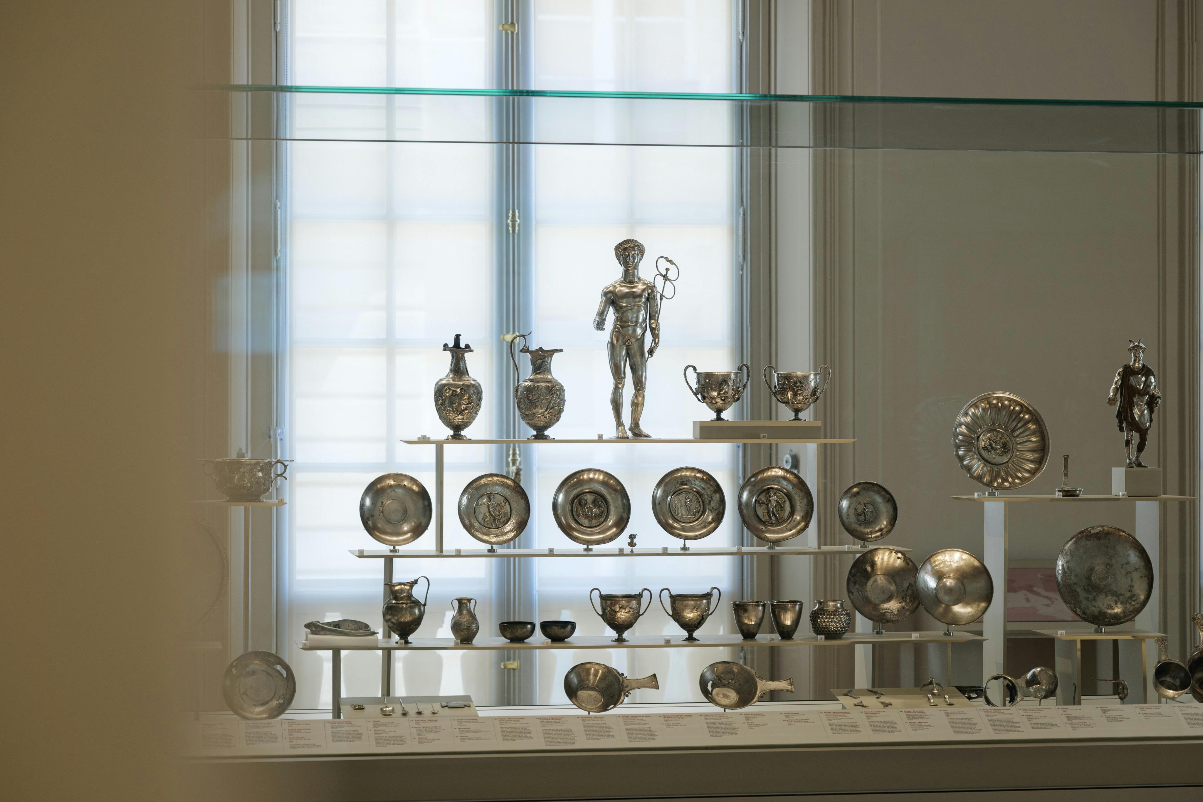 A museum display case showcasing antique silverware, including bowls, cups, and a statue, arranged on shelves.