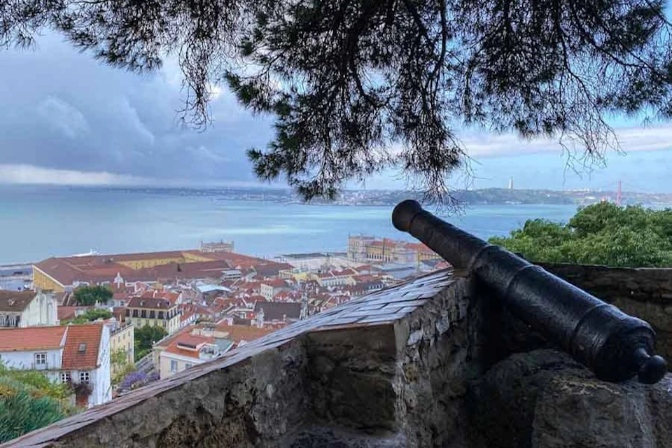 A scenic view of a coastal city with red-roofed buildings, seen from behind an old cannon on a stone fortification.